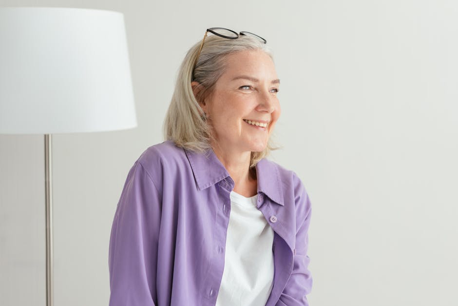 A cheerful senior woman with grey hair wearing a purple shirt smiles indoors.